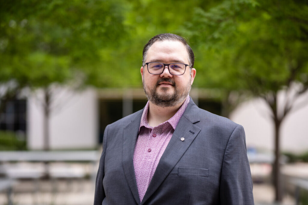A man with a beard and glasses stands outdoors in front of green trees. He is wearing a dark suit jacket over a patterned shirt. The background features a blurred view of a building and benches. The man is looking directly at the camera with a confident expression.