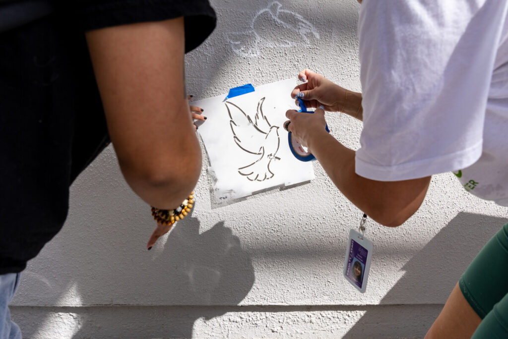 Two individuals are working on a wall, one holding a stencil of a dove while the other is using tape to secure it in place. The person on the left is wearing a black shirt and has a beaded bracelet, while the person on the right is wearing a white shirt and has a lanyard around their neck. The background features a light-colored wall with shadows cast by their hands and the stencil.