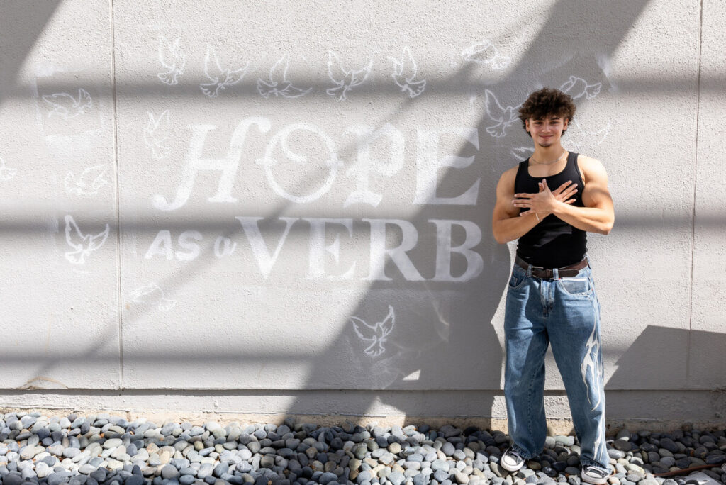 A young person stands confidently against a textured wall, wearing a black tank top and loose-fitting jeans. They have curly hair and are posing with their arms crossed over their chest. Behind them, the wall features the words "HOPE AS a VERB" in large, bold letters, accompanied by white illustrations of doves. The scene is illuminated by natural light, creating shadows that add depth to the image.