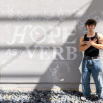 A young person stands confidently against a textured wall, wearing a black tank top and loose-fitting jeans. They have curly hair and are posing with their arms crossed over their chest. Behind them, the wall features the words "HOPE AS a VERB" in large, bold letters, accompanied by white illustrations of doves. The scene is illuminated by natural light, creating shadows that add depth to the image.