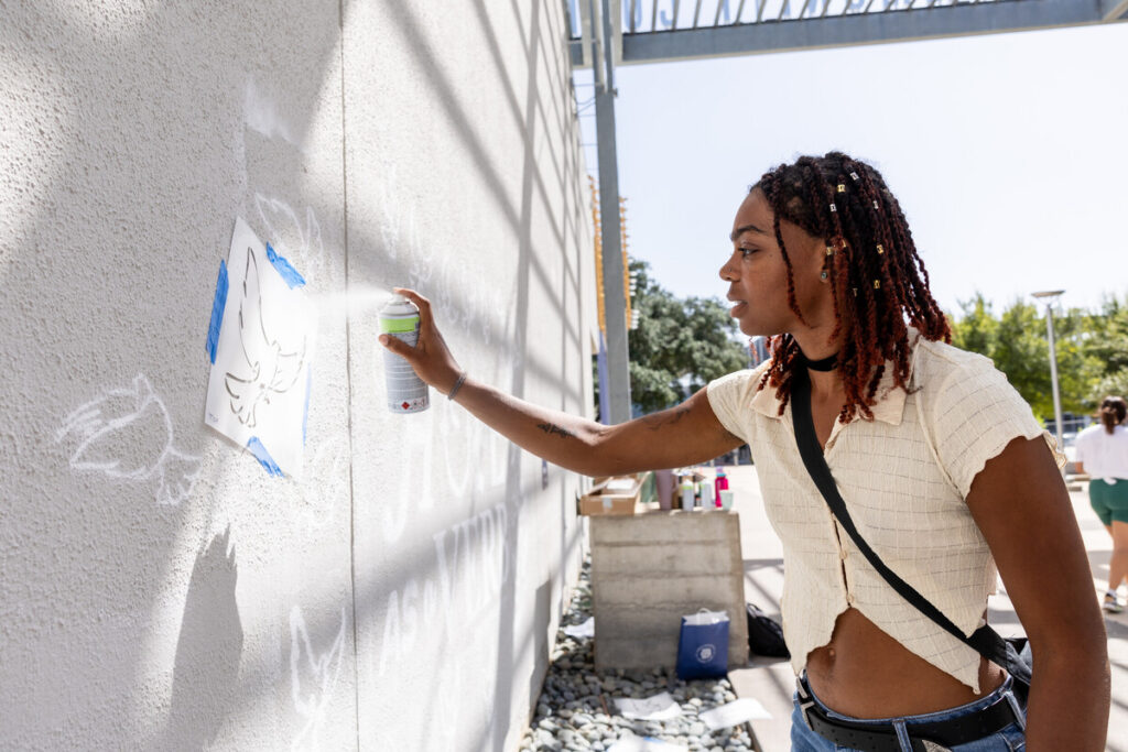 A young person with shoulder-length, braided hair is spray painting a design on a light-colored wall. They are wearing a short-sleeved, fitted top and have a small bag slung over one shoulder. The area around them is bright and sunny, with other people visible in the background, engaged in various activities. The wall has blue tape outlining the area being painted.