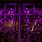 The image features a wall adorned with a pattern of large, three-dimensional stars, illuminated in vibrant purple lighting. The stars are arranged in a grid-like formation, creating a striking visual effect. Behind the stars, soft yellow light emanates from the interior, adding contrast to the overall scene. The architectural structure appears modern and artistic, enhancing the dynamic interplay of light and shadow.