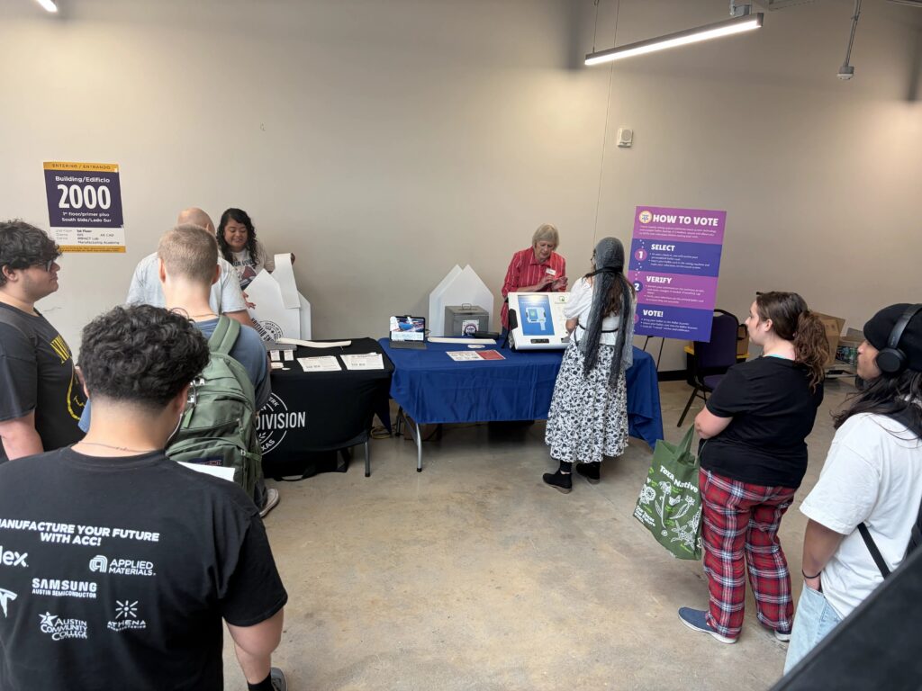 The image shows an indoor voting educational demonstration or voter outreach event set up inside a facility. Voting Station Setup: A table covered in a blue cloth displays voting machines, paper materials, and ballot box equipment, with staff members demonstrating the voting process to attendees.