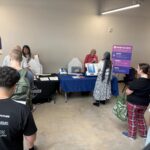 The image shows an indoor voting educational demonstration or voter outreach event set up inside a facility. Voting Station Setup: A table covered in a blue cloth displays voting machines, paper materials, and ballot box equipment, with staff members demonstrating the voting process to attendees.
