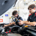 Two automotive technicians are working under the hood of a vehicle in a well-lit garage. One technician, wearing glasses and a black and red uniform, is using a diagnostic tool to check the engine, while the other technician is focused on a different part of the engine. The environment is equipped with various tools and equipment, and a Toyota logo is visible in the background.