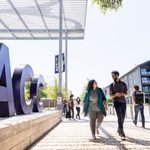 A sunny day on a college campus featuring large, bold letters spelling "ACC" in the foreground. Two students, a woman in a teal jacket and a man in a striped shirt, walk together along a pathway, engaged in conversation. In the background, other students are seen walking, and modern buildings are visible, along with trees providing shade.