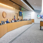 A meeting room with a long wooden table where several individuals are seated, facing a speaker at a podium. The speaker, a woman, is addressing the group, which includes a diverse panel of men and women. The room features a large emblem on the wall, flags, and multiple screens displaying information. The atmosphere appears formal and professional.