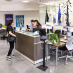 Alt text: A reception area in a military support office. Two women are at the front desk, one assisting a visitor who is standing in front of them. A man with a backpack is waiting nearby, while another man sits at a desk working on a computer. The space features flags and military-themed decorations, with a modern, open layout and plants on the counter.
