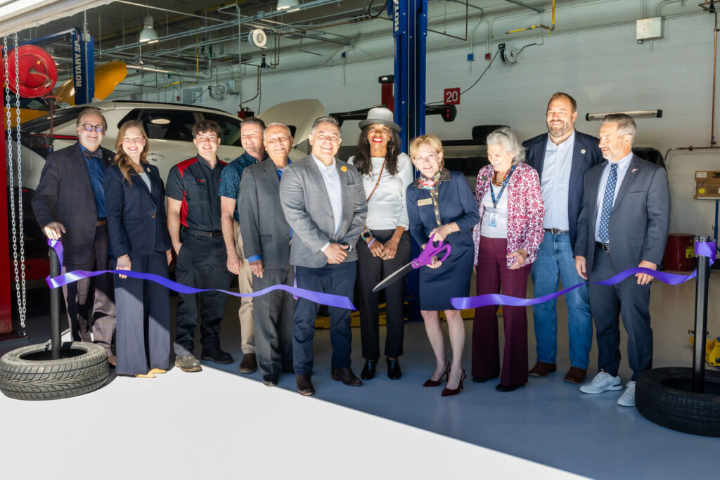 A group of people stands together in a garage-like setting, smiling and posing for a photo. They are gathered around a large purple ribbon, which is about to be cut with oversized scissors by a woman in a white hat and a navy blazer. The group includes men and women of various ages, dressed in formal and casual attire. In the background, there are vehicles and equipment typical of an automotive or aviation workshop. The atmosphere is celebratory, indicating a special event or opening.