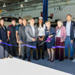 A group of people stands together in a garage-like setting, smiling and posing for a photo. They are gathered around a large purple ribbon, which is about to be cut with oversized scissors by a woman in a white hat and a navy blazer. The group includes men and women of various ages, dressed in formal and casual attire. In the background, there are vehicles and equipment typical of an automotive or aviation workshop. The atmosphere is celebratory, indicating a special event or opening.