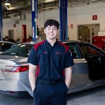 A young man with curly hair stands confidently in a car repair shop, wearing a black and red uniform. Behind him, a silver sedan is parked with its door open, and various tools and equipment are visible in the background, indicating an active automotive service environment.
