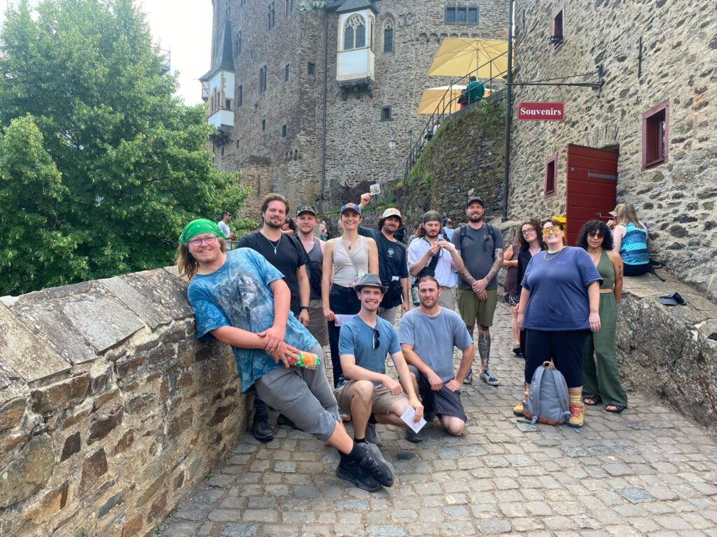 The backdrop features a tall stone fortress wall with an ornate window balcony, an open red doorway, and a red "Souvenirs" sign fixed to the masonry in Germnay. A group of about a dozen young adults are gathered together, including one person in a green bandana and blue tie-dye shirt leaning back against the left retaining wall, two individuals seated or kneeling in the front, and others standing along the walkway. An elevated outdoor terrace with patio umbrellas and dining tables is visible higher up along the castle structure