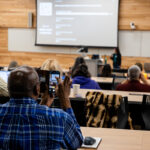 A classroom setting with a diverse group of students seated at desks, facing a presenter at the front. The presenter is standing next to a screen displaying information, while a student in the foreground, wearing a blue plaid shirt, is taking a photo with a smartphone. The room features wooden paneling and modern furnishings, creating a professional learning environment.